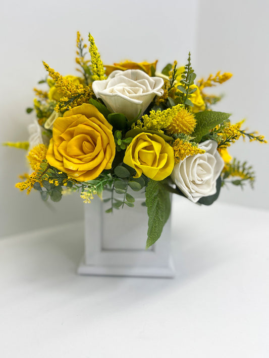 Yellow & ivory handcrafted wooden roses with greenery placed in white, square planter. 