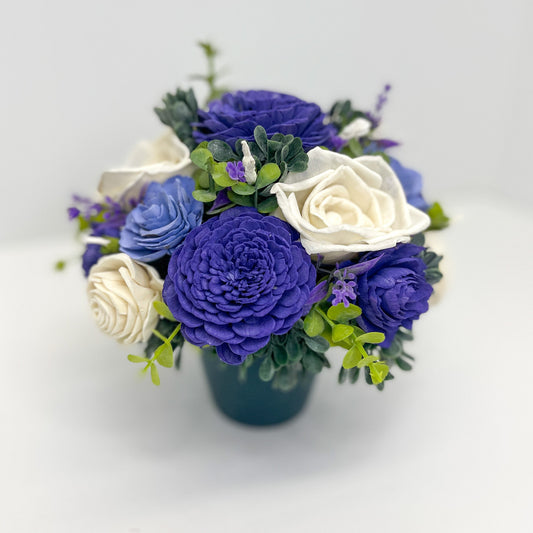 Dark purple, light purple & white wooden flower bouquet with greenery placed in planter. 