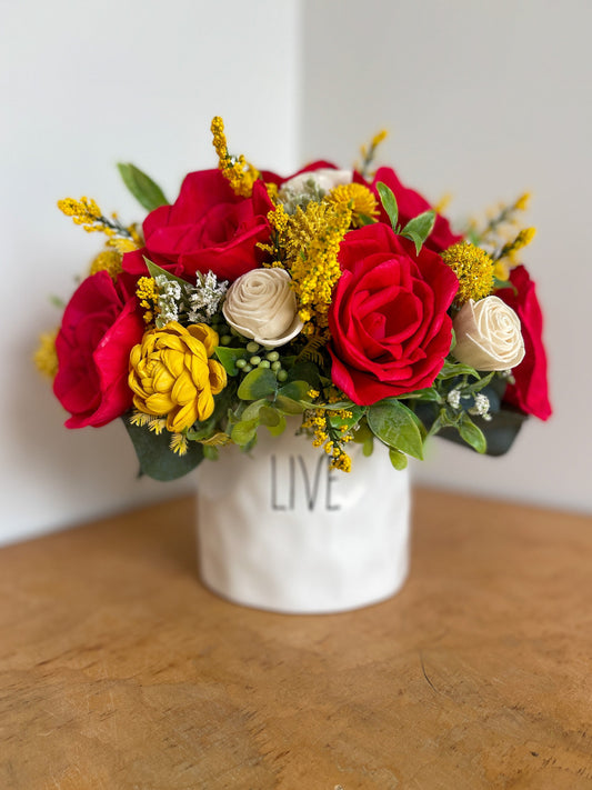 Red, yellow & white wooden flower bouquet with greenery placed in white planter with "LIVE" in black print. 