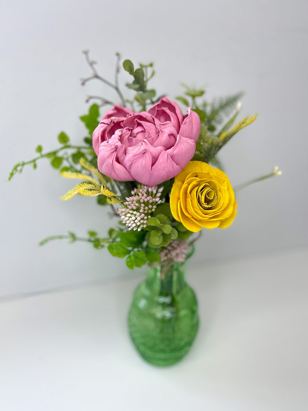 Light pink and yellow wooden flowers with greenery placed in translucent green bud vase. 