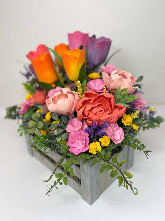 Bright colored bouquet with pink, purple & yellow wooden flowers and greenery placed in grey crate. 