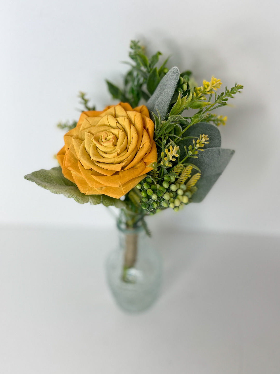 Single stem yellow wooden flower bouqet with greenery placed in clear bud vase. 