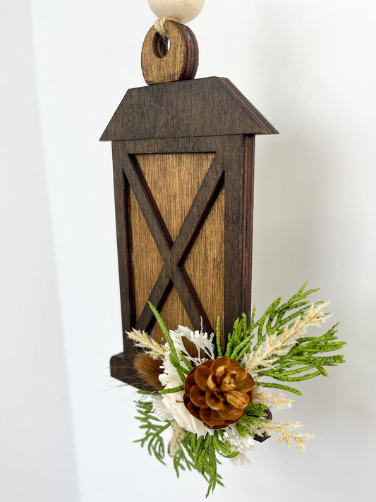 Dark and light brown wooden lantern ornament decorated with wooden pinecone and pine needles.